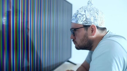 Man wearing an aluminum foil hat expresses disbelief while watching a vibrating screen in a humorous conspiracy scene