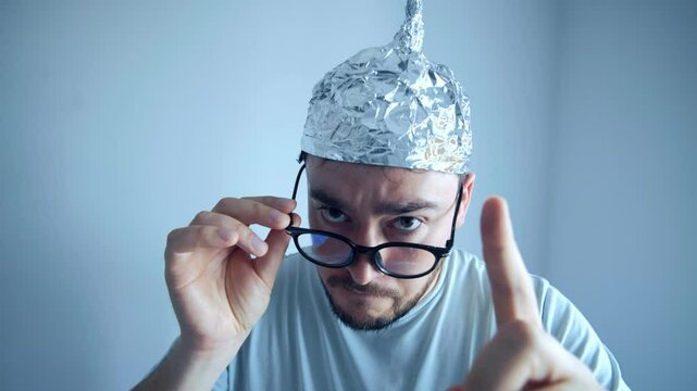 Man wearing a tinfoil hat displays paranoia and disbelief while adjusting his glasses in a humorous setting