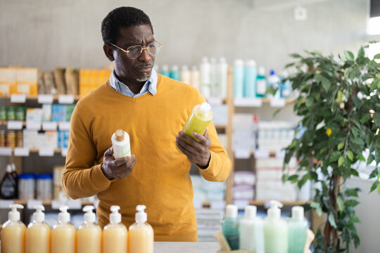 African man choose buy antidandruff shampoo at pharmacy, compare with other good. Client check for absence of harmful substances, view product in details.