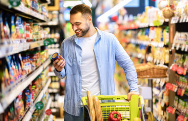 Consumption Concept. Portrait Of Smiling Bearded Guy With Shopping Cart In Market Buying Groceries Food Walking Along The Aisle And Shelves In Store, Holding Glass Jar Of Sauce, Checking Label