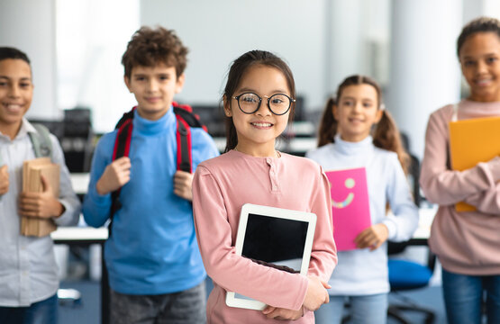 Pupils, Technology And Learning Concept. Smiling asian girl in eyeglasses holding digital tablet and standing with group of multicultural classmates in the background, studying at elementary school