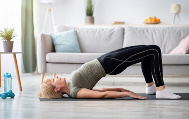 Stay at home fitness. Strong mature woman doing half bridge yoga pose, strengthening her abs muscles indoors, copy space. Fit senior woman working out in living room during covid isolation