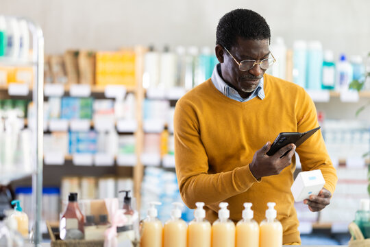 Focused black man in fifties in pharmacy scanning barcode on box of pills with smartphone, carefully checking dosage, side effects, or contraindications before purchase, ensuring safe self-medication