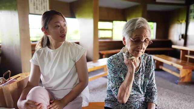 A sunny early summer day at a canal park in Toyama City. A Japanese grandmother in her 90s and a woman in her 30s are sitting together at a rest area, chatting happily and enjoying the peaceful time.