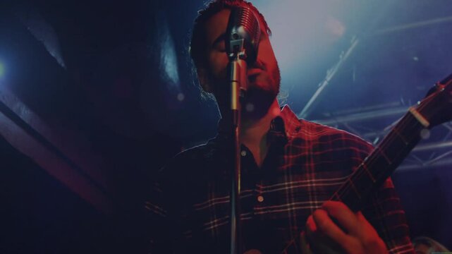 Music industry musician singing into vintage microphone strumming guitar under colored spotlights