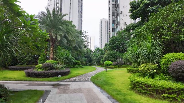 A lush tropical communal garden with a walking path, manicured lawns, ornamental shrubs, and trees in a shared communal courtyard between residential apartment buildings in China.
