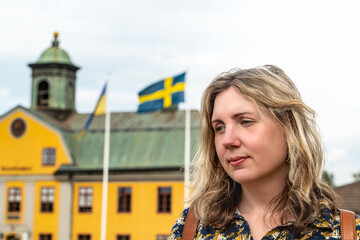 A woman poses thoughtfully with the Swedish and Ukrainian flags in view, set against a colorful building in Sweden during a sunny summer day