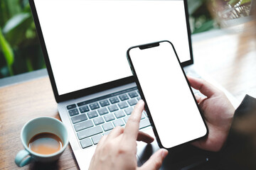 Person pointing at smartphone with blank screen while working on laptop at a cafe with a cup of coffee.