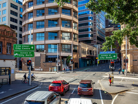 Elevated view in Sydney, Australia downtown intersection featuring modern office buildings, directional signs, vehicles, and pedestrians waiting at a traffic light in an urban business district.