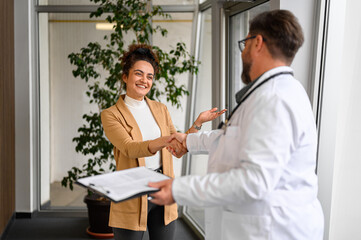 Male doctor in lab coat shaking hands with female patient while standing in hospital during medical visit