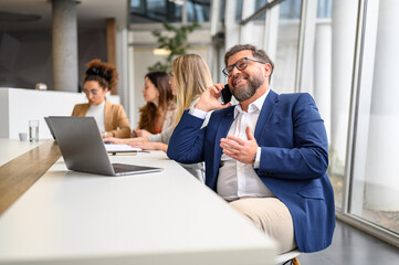 Businessman with laptop talking on smartphone while female colleagues working in the background in office