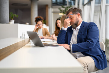 Stressed businessman rubbing his eyes while working over laptop with females colleagues in the...