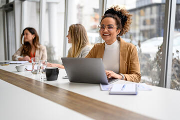 Happy businesswoman in eyeglasses working over laptop while siting with colleagues in modern board room