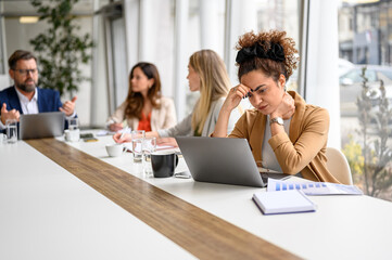 Stressed businesswoman focusing on her laptop while colleagues discussing and collaborating in the background during meeting