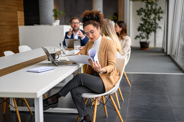 Businesswoman reviewing reports and working over laptop with colleagues in the background at modern...