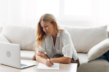 Woman working from home, writing notes on paper while sitting on a comfortable couch with a laptop nearby