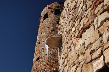 Close-up of a rustic stone tower with icicles hanging from a ledge against a deep blue sky in winter sunlight..