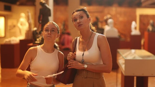  Young woman with teen daughter walks around museum hall, examines showpiece sculpture at exhibition. List of recommended exhibitions, guidebook, and map of museum in hands