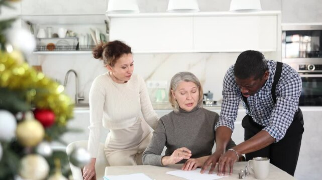 Thoughtful old woman compiling testament while middle-aged man and woman competing with each other in the kitchen decorated with Xmas-tree. High quality 4k footage