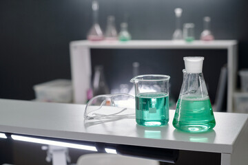 Laboratory counter showcasing diverse scientific equipment with beakers containing green chemical solutions, safety glasses visible in background