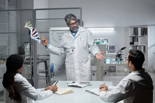 Scientist presenting DNA model while standing in front of transparent board, engaging with colleagues seated at table. Modern laboratory visible in background with various scientific equipment
