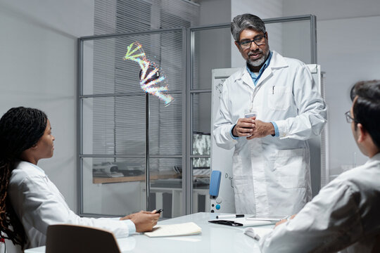 Male scientist explaining genetic code to colleagues in lab setting with transparent display showing 3D DNA model. Discussion engaging researchers around central table