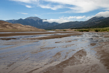 River gently flows through sandy landscape under cloudy mountain skies in Great Sand Dunes National Park