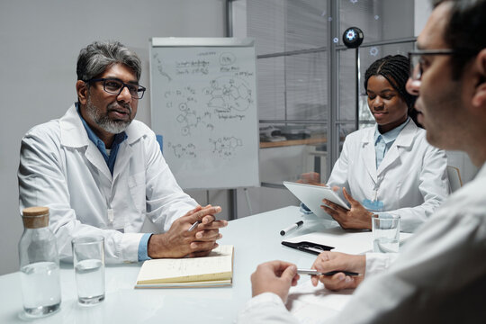 Diverse group of scientists engaging in collaborative discussion in modern laboratory environment with scientific equipment and whiteboard filled with chemical equations in background