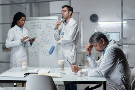 Group of scientists in lab coats sharing ideas and reviewing notes while standing near a whiteboard and table filled with research materials, coffee cups, and equipment