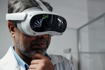Middle-aged man in laboratory environment examining virtual content displayed in advanced VR headset thinking deeply wearing white lab coat. Modern technology in scientific research