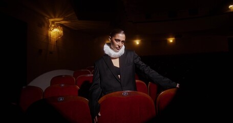 A woman with a frill around her neck walks between the chairs in an empty auditorium in a theater....