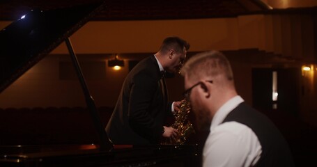 A stylish man plays the saxophone while standing on stage. The second man is playing the piano