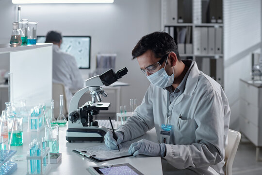Scientist wearing protective gear and a face mask, recording observations in lab. Various scientific equipment and colorful chemical samples visible in workspace