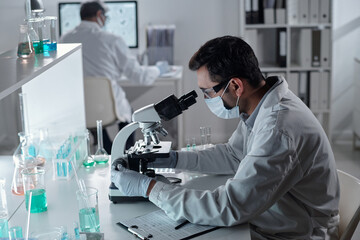 Scientist wearing lab coat and protective mask analyzing sample with microscope in laboratory...