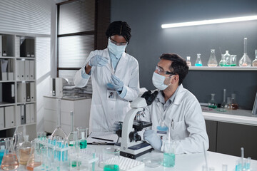 Two scientists wearing masks are working in a laboratory performing analysis with microscope and beakers on the table, communicating and focusing on scientific research
