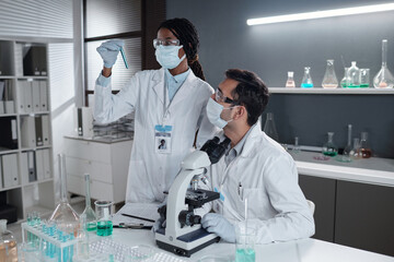 Scientist examining blue liquid in test tube while colleague observing through microscope in modern laboratory setting involving scientific research and analysis