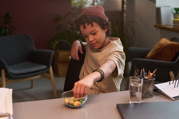 African American schoolchild taking candy from glass bowl while sitting by workplace of school psychologist in his office