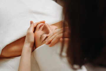 face massage, young attractive caucasian woman, beautiful relaxed lady with closed eyes smiling, white towel, spa therapist doing facial and head massage in beauty salon, skincare and bodycare concept