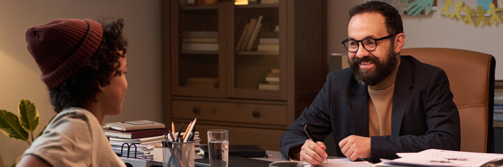 Header of happy mature school psychologist looking at African American boy sitting in front of him during discussion of issue