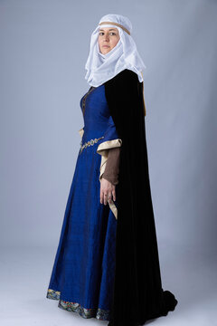 A Medieval woman wearing a blue silk gown called a bliaut and a white linen wimple against a plain studio backdrop