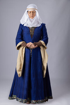 A Medieval woman wearing a blue silk gown called a bliaut and a white linen wimple against a plain studio backdrop