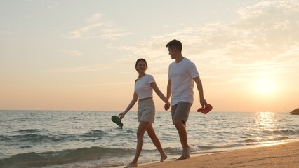 Fototapeta premium Vacation loving couple walking on beach together at sunset landscape, relaxing vacation travel summer, Asian man and woman in love holding hands walks along the sandy seashore