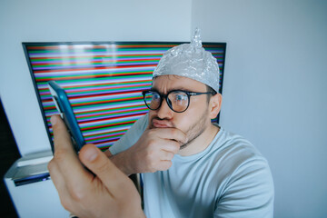 Man with aluminum foil hat displays fear and disbelief while checking phone for conspiracy theories...