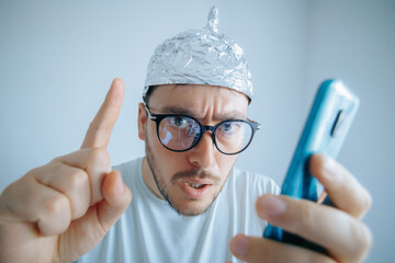 Man in aluminum foil hat expresses disbelief while holding smartphone in room