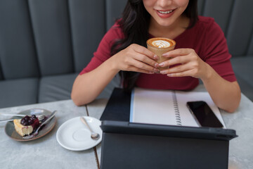 Asian freelancer woman is smiling while holding a coffee cup, working on a tablet in a cafe with a notebook, smartphone, and cheesecake on the table