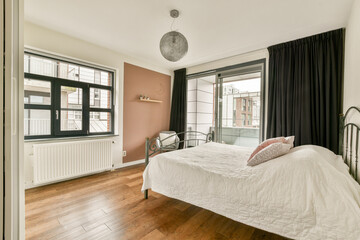 A stylish bedroom featuring a bed with white bedding, large windows allowing sunlight, and a warm wooden floor, creating a serene atmosphere.