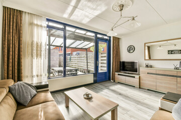 A stylish living room featuring large windows, a comfortable sofa, and wooden furniture. The decor balances modern and cozy elements, creating an inviting atmosphere.