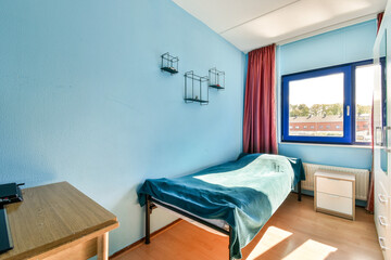 A well-lit and inviting room featuring a blue wall, a large window with curtains, and simple furniture, creating a pleasant living space.