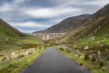 A charming pathway gently winds through an inviting, vibrant Silent Valley Reservoir, showcasing a substantial dam nestled among the rolling green hills beneath a tranquil cloudy sky, Northern Ireland