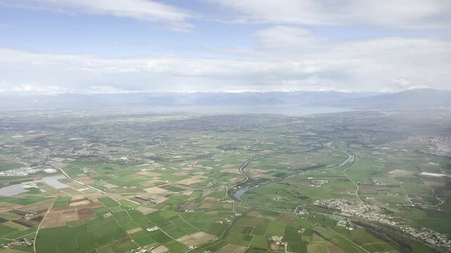 High-altitude aerial footage flying over the colorful farmland of Veneto region with Adige River winding towards Lake Garda, snowy peaks of the Alps visible under a partly cloudy sky